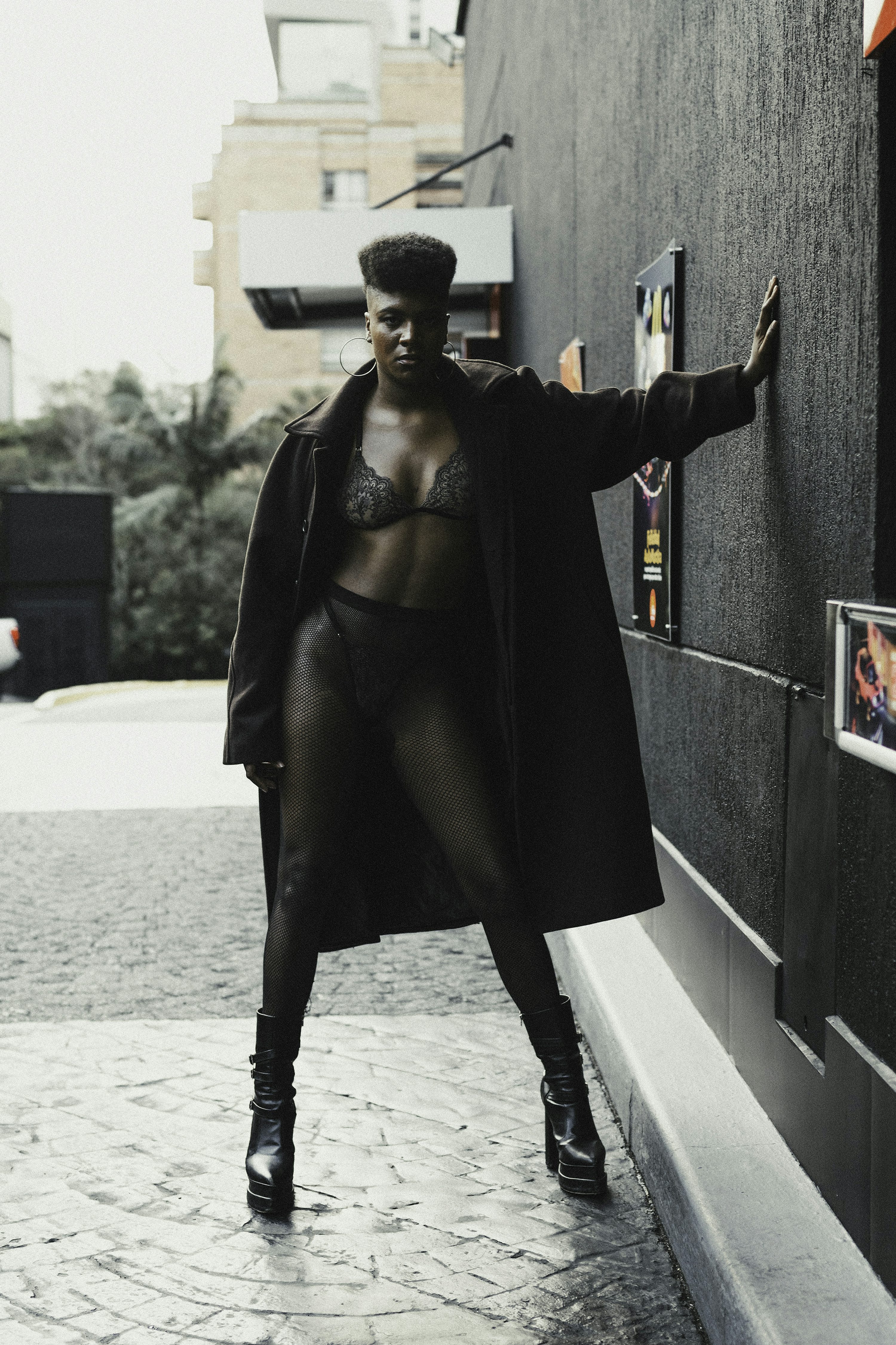 a woman with short hair is posing outdoors against a dark wall. she is wearing a long dark coat over lingerie and black highheeled boots.