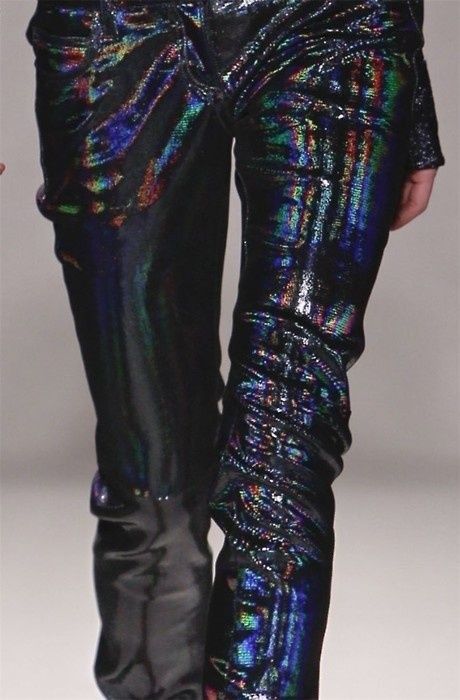 a closeup shot of a model wearing a pair of black iridescent pants on a runway. the pants are shiny and have a sequined look. the model's legs are covered in the pants, showing off the detail of the fabric.