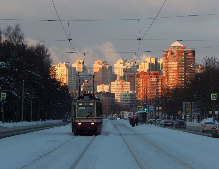 a photograph of a tram traveling down a snowcovered street in a city. the street is lined with trees and highrise residential buildings. the sun is setting, casting a warm glow on the buildings. the image is taken from a perspective that shows a section of the street including the tram tracks, people, cars, and tall buildings in the background.  the overall feeling is of a quiet, cold winter evening in an urban setting.