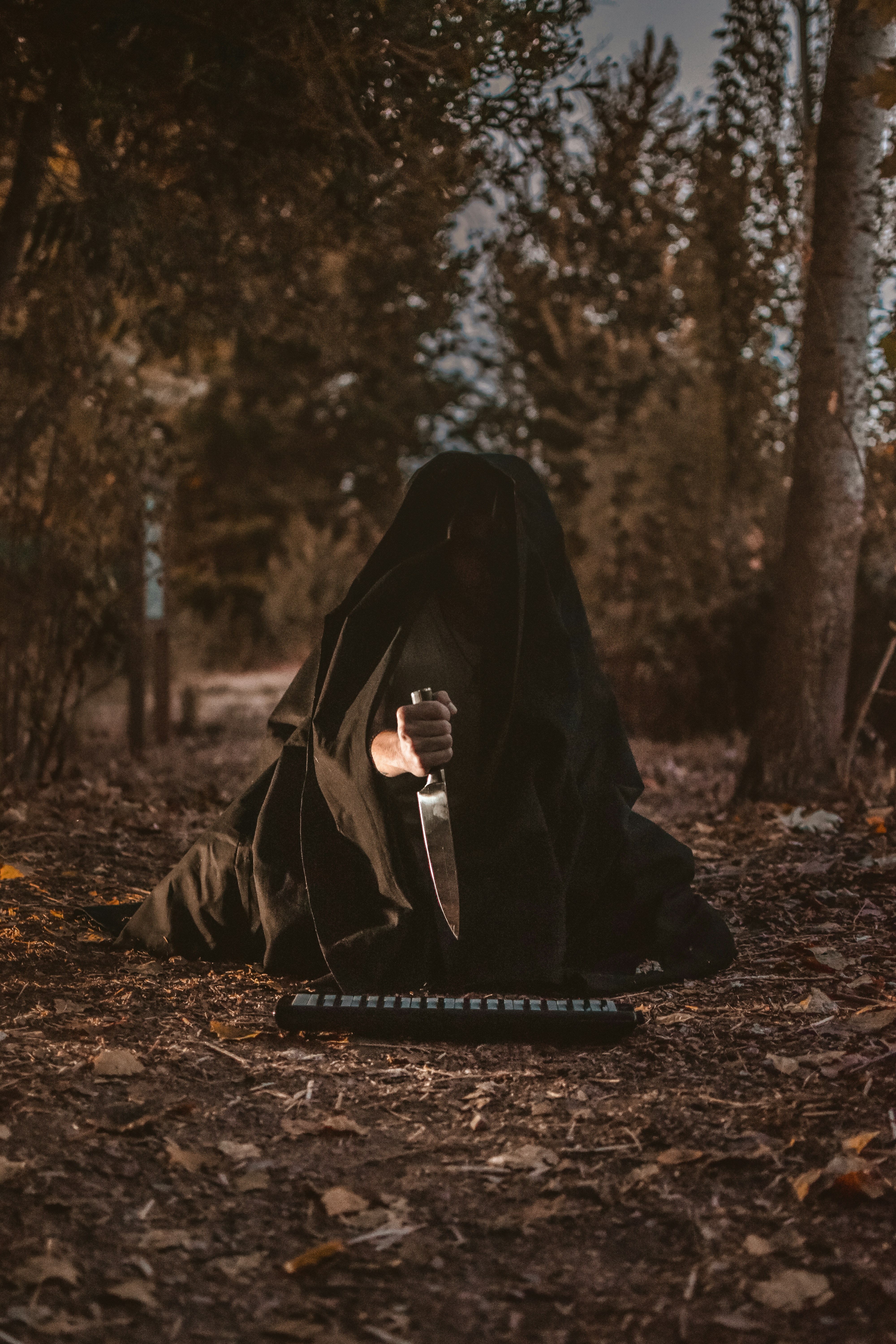 a figure draped in a black cloak sits hunched on a forest path, clutching a large knife. the figure's face is hidden within the depths of the hood, obscuring their identity. the setting is a dimly lit forest, likely at dusk, with warm sunlight filtering through the trees. the overall atmosphere is ominous and suspenseful, hinting at a potential threat.