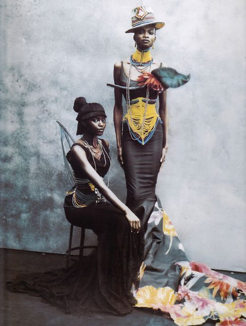 two fashion models posed in a studio setting, wearing ornate, black clothing, necklaces and bracelets, and floral headwear. one model sits on a chair, wearing a long black dress with a train, while the other model stands, wearing a fitted black dress with a long flowing floral train. they both strike elegant poses for the camera.  the background is gray, adding to the dramatic effect of the models' poses and clothing.