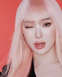 the image is a closeup portrait of a young woman, likely a kpop idol, with long, light pink or pastel blonde hair styled with bangs. she's winking at the camera, and her makeup is soft and subtle. the background is a solid, bright coral or reddishpink. she's wearing what appears to be a darkcolored top. the overall aesthetic is very soft, cute, and stylish, common in kpop promotional imagery.