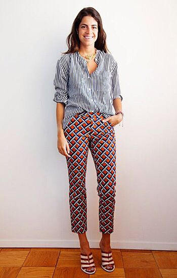 a fullbody shot of a stylish woman posing indoors against a white wall. she is wearing a navy and white striped longsleeve shirt, cropped geometric print pants in orange, navy and white, and brown and white striped sandals. she is wearing a gold necklace. her hair is long and brunette. the floor is hardwood. the overall style is casual and chic.