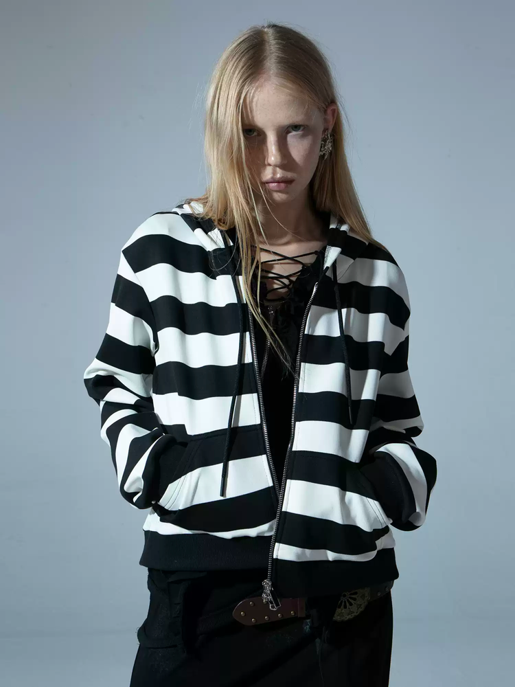 a blonde woman is posing in a black and white striped zipup hoodie. she is wearing dark pants and a black top underneath. the hoodie has a relaxed fit and features prominent black and white stripes. the model is looking directly at the camera. the image is a studio shot, with a plain background.