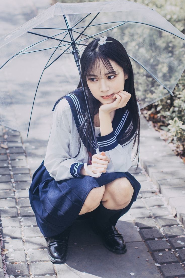 a young asian woman in a school uniform, consisting of a white shirt with a sailor collar and a dark blue skirt, is crouching on a cobblestone path. she is holding a clear umbrella and appears pensive, resting her chin on her hand. the background is blurred, suggesting an outdoor setting. she is wearing black shoes and knee socks.