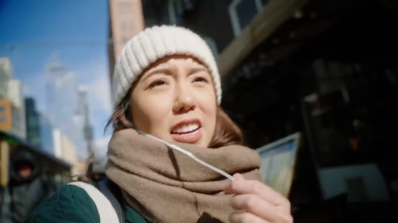 a young asian woman takes a selfie while exploring a bustling city street.  the sun is shining brightly, illuminating her face and the surrounding buildings.  she's bundled up in winter clothes including a white beanie and brown scarf, and seems to be enjoying her time sightseeing.