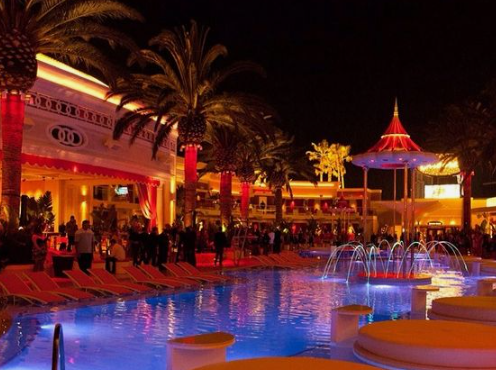 an outdoor pool area at night, filled with people enjoying a party. the scene is lit with vibrant neon lights in red, blue, and white, illuminating the pool, palm trees, surrounding buildings, and a central gazebo structure. people are socializing, standing and sitting on red and white lounge chairs surrounding the pool. the overall ambiance is festive, luxurious, and energetic.