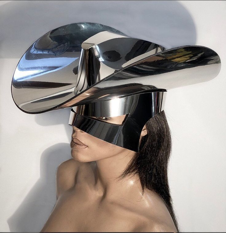 a woman wearing a futuristic silver hat and mask. the hat is shaped like a cowboy hat with a visor and the mask covers her eyes. the woman has long dark hair and is looking to the side. the image is in color and is taken from a closeup perspective.  the model is wearing a silver hat and mask, with a bright white background.  the hat and mask are a unique design, with sharp edges and a sleek finish. the model's hair is long and dark, and her face is partly obscured by the mask. the image is striking and eyecatching, and it captures the model's beauty and style.  the image is a beautiful example of fashion photography, and it showcases the model's unique style.