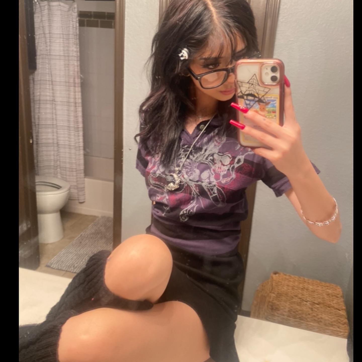 a young woman with dark hair and glasses takes a mirror selfie in a bathroom. she is wearing a purple graphic tshirt, a black skirt, and black leg warmers. she has red nails and is wearing a silver necklace and bracelet. the bathroom has a white toilet and a beige shower curtain. the lighting is artificial and the perspective is from her point of view in the mirror.