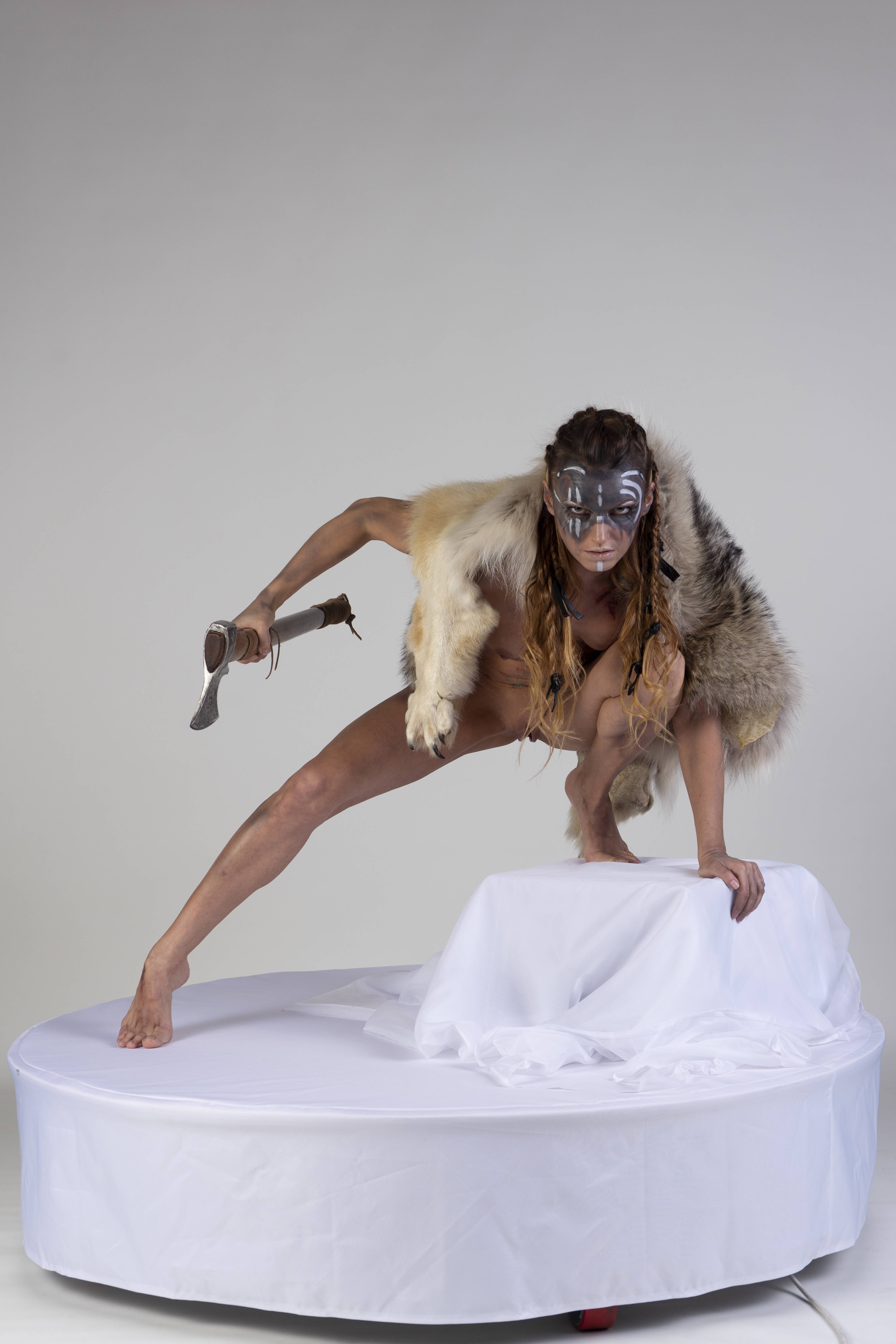 a warrior woman, adorned with body paint and a fur garment, crouches aggressively on a white platform while wielding an axe. she is barefoot and appears to be in a studio setting.