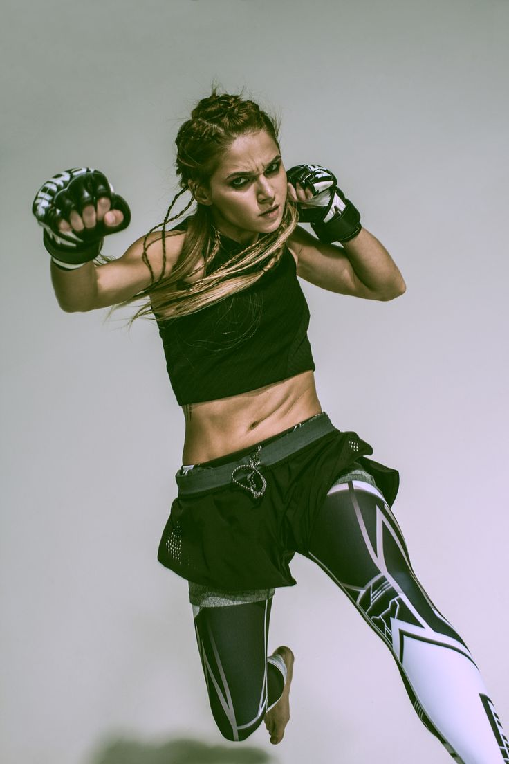 female athlete with braided hair is captured midair, executing a powerful kick.  she's dressed in black and grey athletic wear and mma gloves, showcasing intensity and focus in her expression. the studio setting and bright, frontal lighting emphasize her dynamic movement. the low angle perspective adds to the image's powerful and energetic feel.