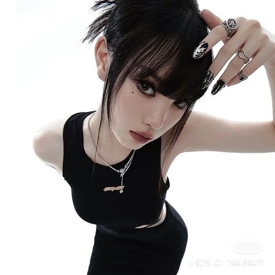 a woman with black hair styled in a messy updo, wearing a black sleeveless dress and silver jewelry, poses against a white background. she gazes intensely at the camera from a low angle perspective. her makeup is minimal and naturallooking, and she has black nail polish with silver accents. the lighting is bright and focused, creating sharp contrasts and highlighting the details of her features and clothing.