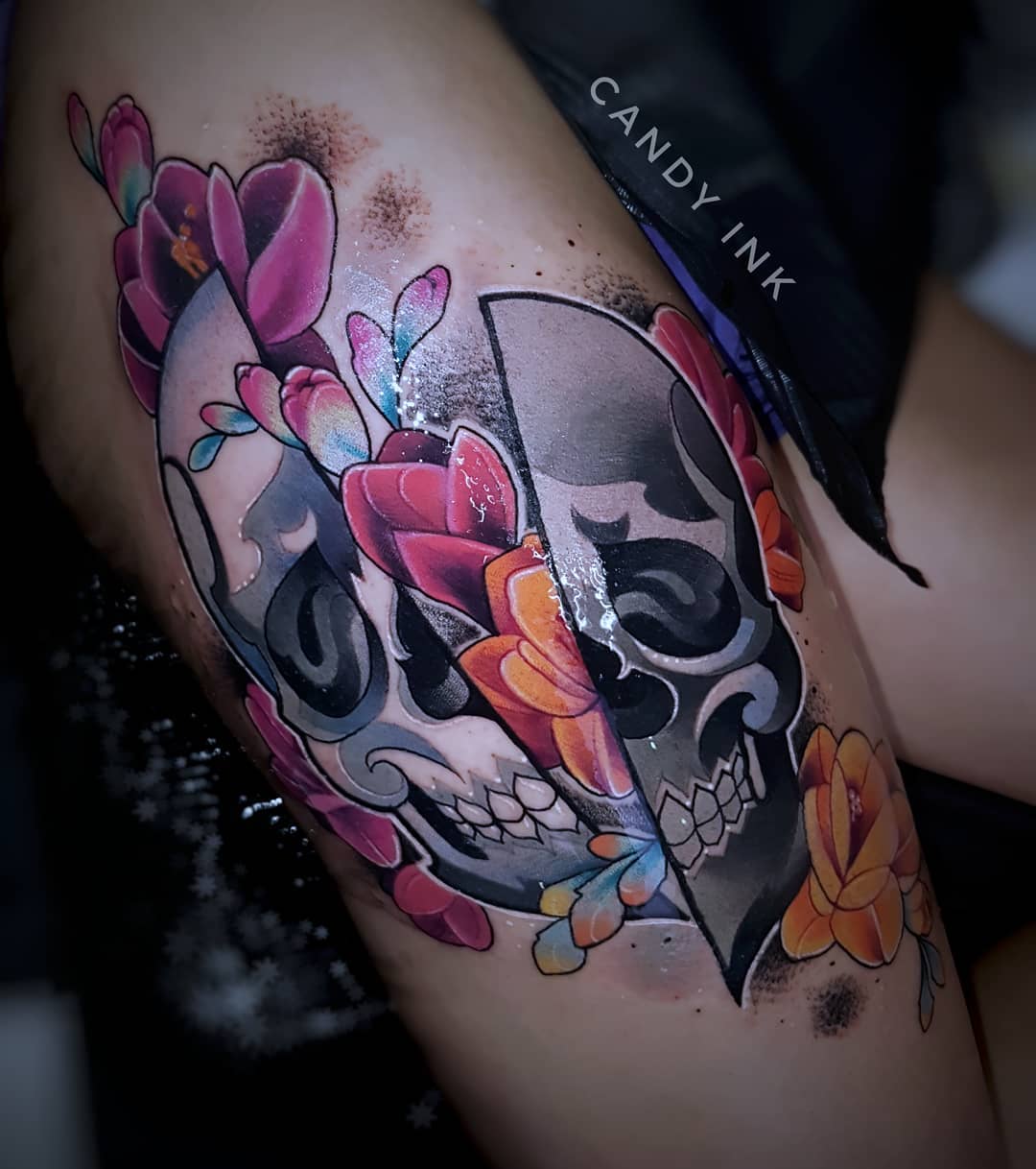 a person is getting a tattoo on their upper leg. the tattoo is of a skull that is split down the middle. one half of the skull is black and white, and the other half is in color. there are flowers surrounding the skull.