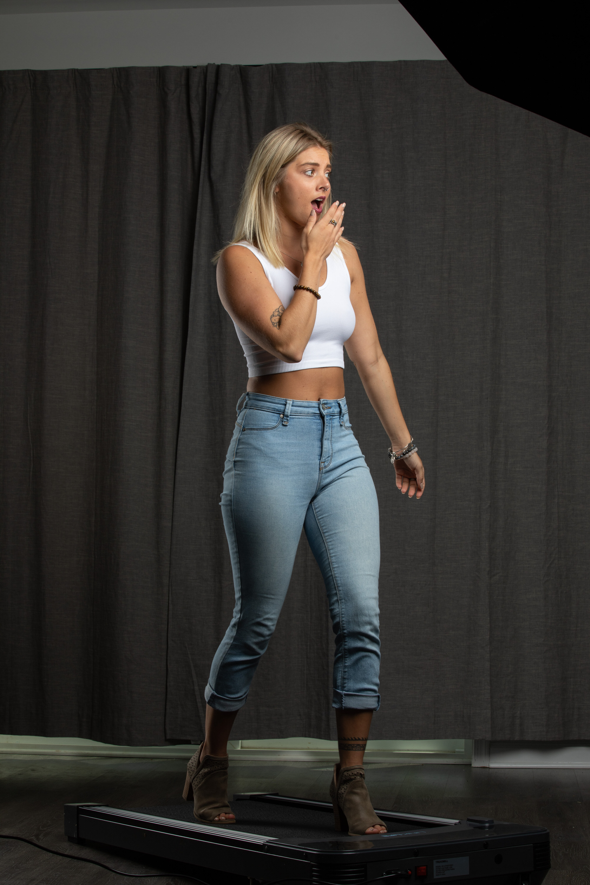 a young blonde woman stands on a treadmill in a room. she's dressed in workout attire with a white crop top and blue jeans, her hand covering her mouth in surprise. her pose suggests she might have just stepped off the machine. the room is simple, with a grey curtain backdrop and a dark grey floor. the lighting is artificial, likely from a studio setup.