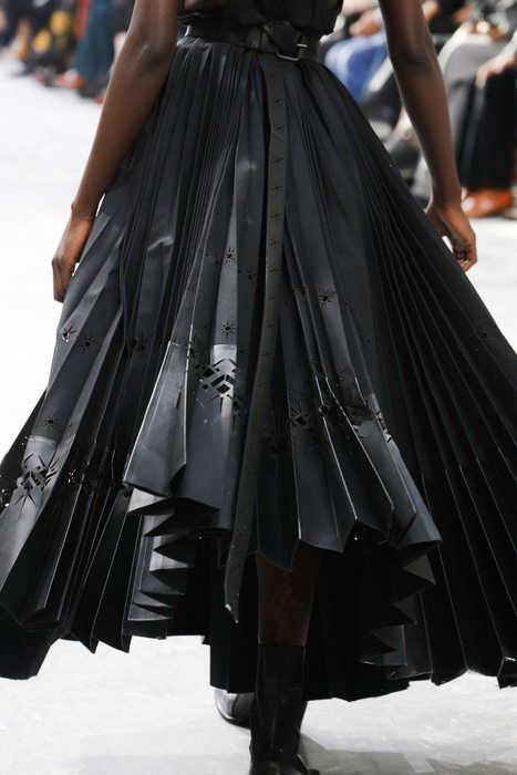 a woman is walking on a runway, wearing a black dress with a heavily pleated skirt. the skirt has geometric cutouts. she is wearing a belt and boots. the background is out of focus, showing other people and the runway setting.