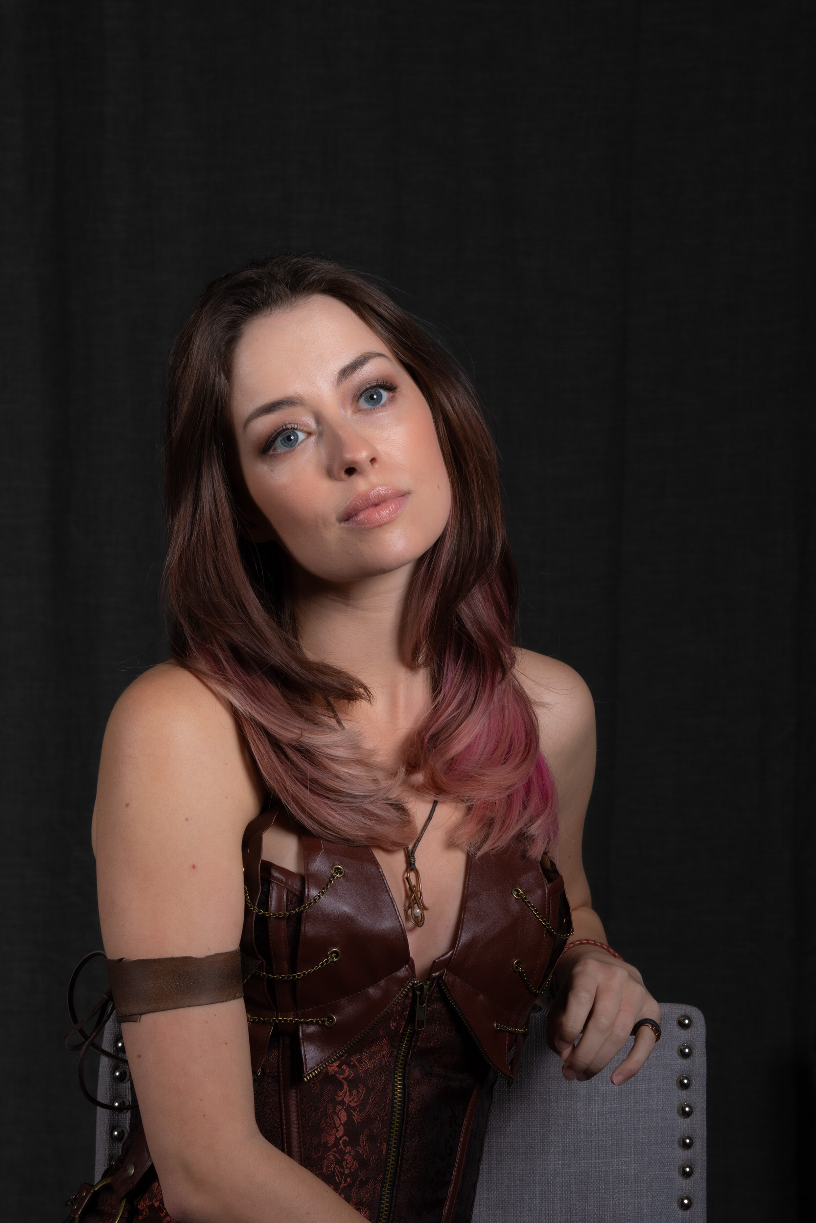 a young woman with long, brown hair streaked with pink sits slightly turned in a chair. she wears a brown, leatherlike corset top and stares directly at the camera. the background is a dark, solid color.