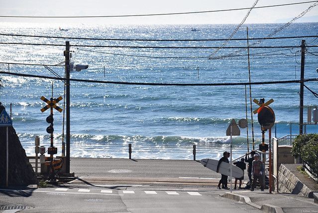 the image shows a picturesque coastal scene in japan. in the foreground is a road crossing with railroad crossing signals.  several surfers are walking along the road, carrying surfboards, having apparently just left the beach. the background features a calm, blue ocean with gentle waves lapping the shore. power lines stretch across the scene, adding to the everyday feel of the setting.  the overall ambiance is serene and peaceful.