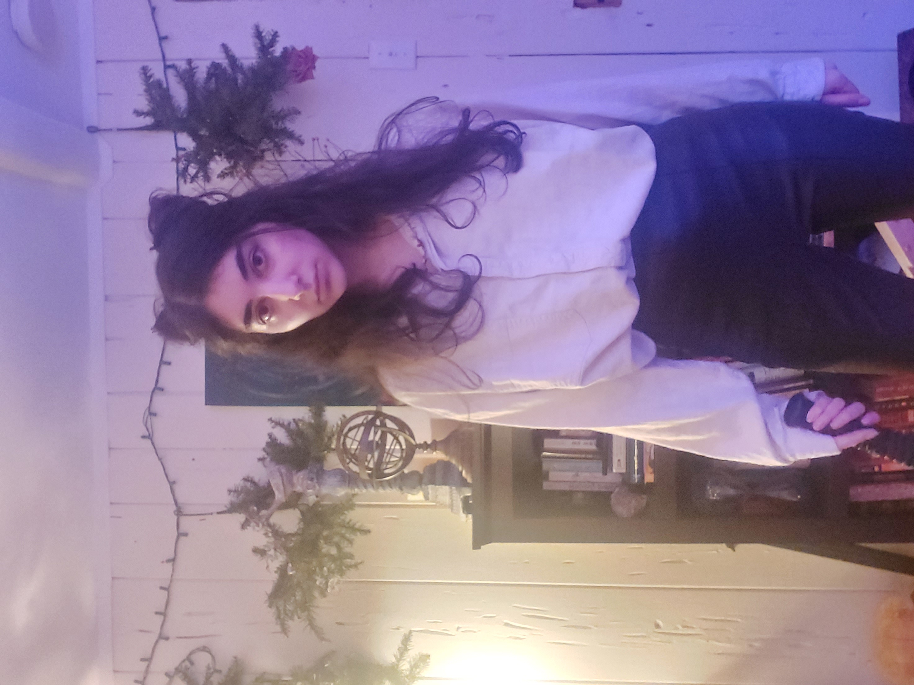 the image shows a young woman, possibly a teenager, standing indoors in what appears to be her home. she's dressed in a light purple, loosefitting buttondown shirt and dark, possibly leather, pants or leggings. her hair is styled in two topknots. the background features a bookshelf filled with books, an armillary sphere, and some christmas lights and evergreen branches hung as decoration. the lighting in the photo is dim, with a predominantly purple hue, creating a moody and atmospheric scene. the young woman looks directly at the camera with a somewhat serious expression.  she is holding a darkcolored object in her hand, but it is not clearly identifiable.  the overall aesthetic is somewhat mysterious.