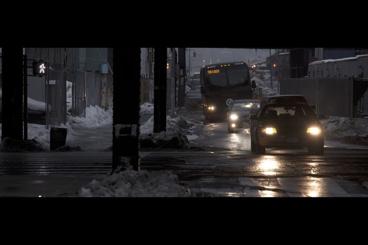 a lowlight image depicts a street scene at night in a city during winter. a large bus is the main focus, followed by several cars driving in the same direction. the street is partially covered in snow, and there's a pedestrian crossing visible in the background. the overall atmosphere is dark, cold, and somewhat gloomy, emphasizing the urban winter setting. the image's depth of field is shallow, with the cars slightly out of focus, drawing attention to the bus.  the headlights of the vehicles illuminate the wet road surface.