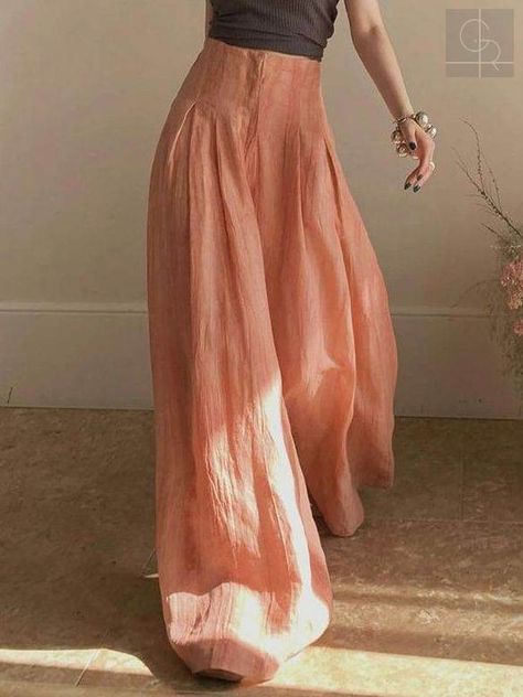 a woman is standing indoors, modeling a pair of highwaisted, wideleg pants. the pants are a peachypink color and have a flowing, loose fit. she is wearing a black sleeveless top and has her arms raised slightly. the image is lit with soft, natural light, creating a warm and inviting atmosphere.