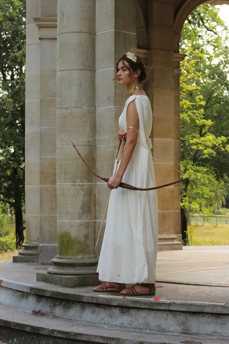 a young woman dressed in a flowing white ancient greekstyle dress stands beneath a stone archway. she holds a bow and arrow, portraying a goddess or huntress. the setting is outdoors, in a parklike area with lush green trees in the background.  the stone architecture is old and weathered, adding to the classical aesthetic.  she is wearing sandals and gold jewelry, including a headband. the overall mood is serene and evocative of ancient mythology.