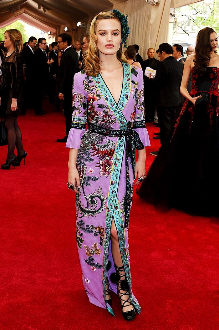 a full shot of a woman in a long, purple, floral embroidered wrap dress at the met gala red carpet. the dress has a kimonostyle silhouette and teal accents. she is wearing black laceup heels and has her hair styled in loose waves.  the red carpet is visible in the background, along with other people attending the event, some partially visible.