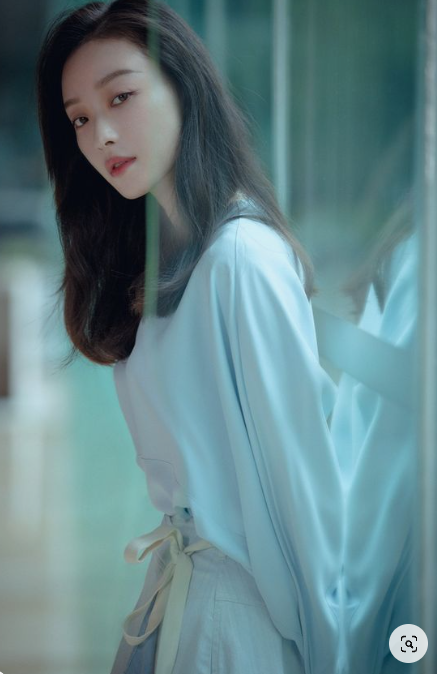 a young woman with long black hair is posing indoors, leaning against a windowpane. she's wearing a stylish, loosefitting pale blue shirt with a bow tied at her waist and a matching pale blue skirt. the lighting is soft and natural, giving the image a serene and elegant feel.