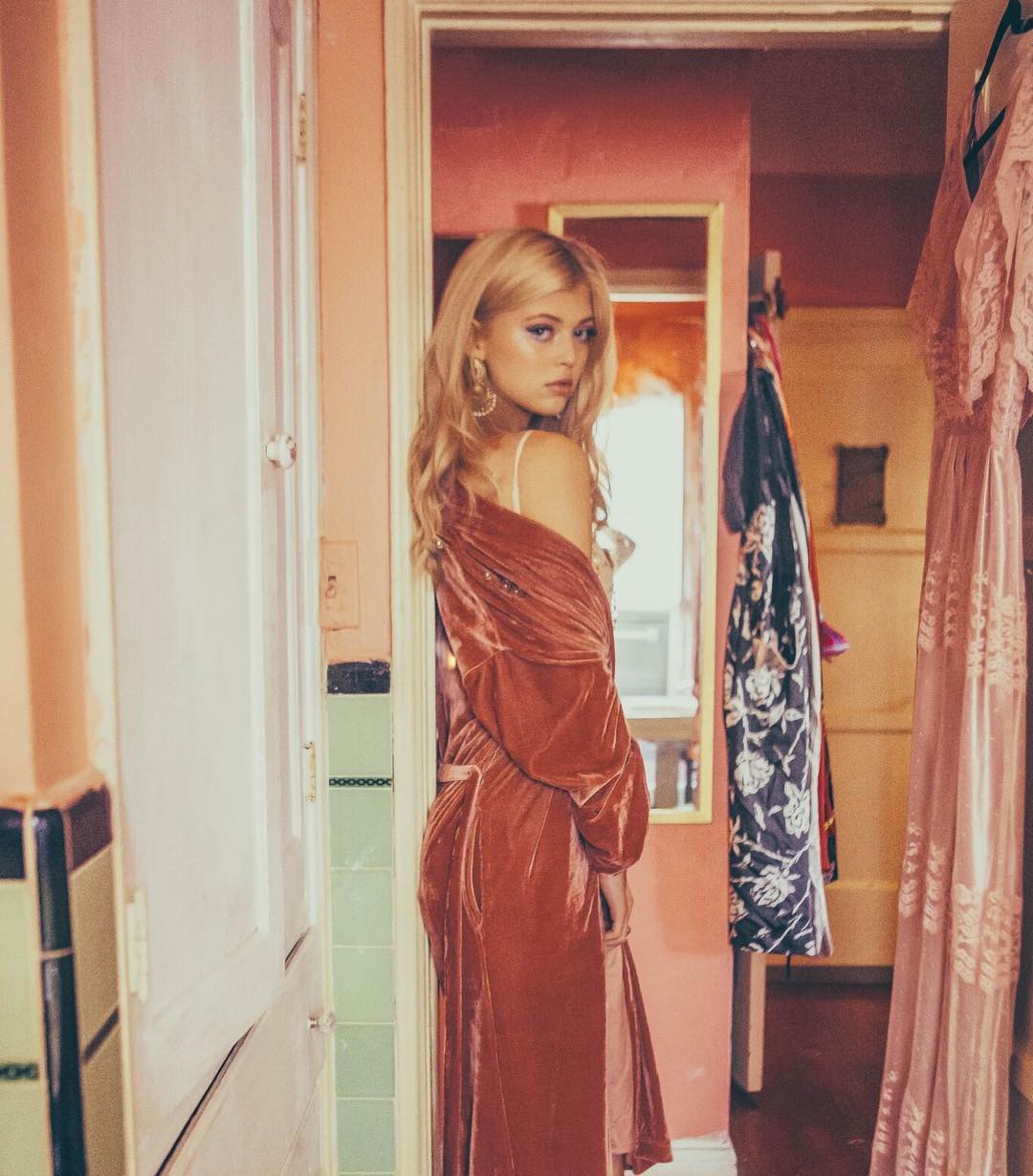 a woman with blonde hair wearing a pink velvet robe stands in a doorway. there is a mirror behind her and clothes hanging in the background.
