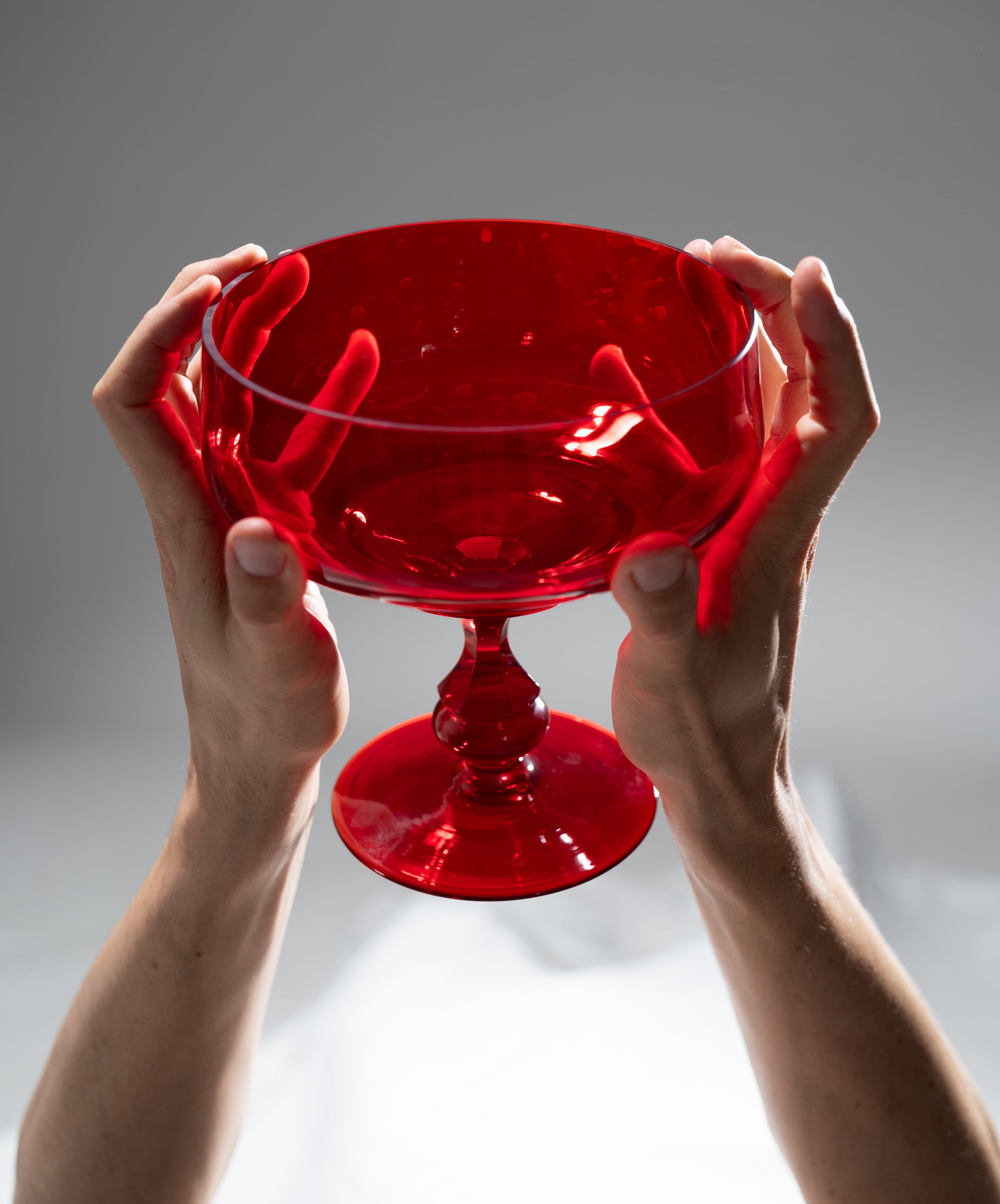 a pair of hands delicately holds a vibrant red glass bowl, illuminated by direct sidelighting against a muted gray backdrop. the glass exhibits a translucent quality, revealing subtle texture and depth in its rich crimson hue.