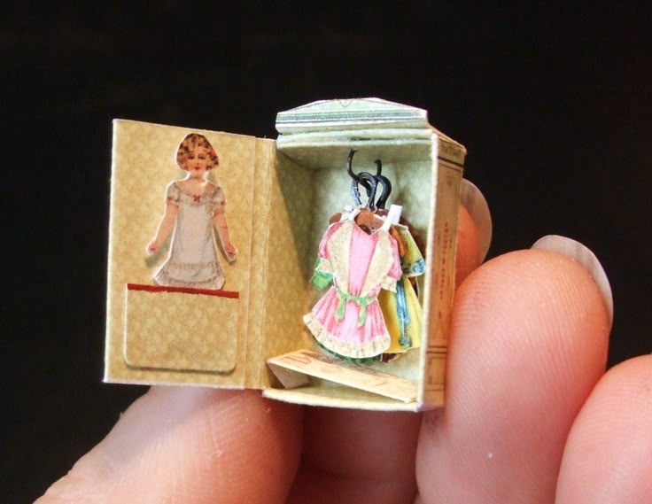 a closeup shot of a miniature wardrobe held between someone's fingers. the wardrobe is open, revealing several tiny dresses on hangers and a paper doll figure on the inside of the door. the wardrobe is intricately crafted with small details, showcasing miniature artistry.