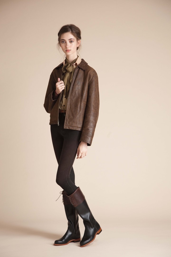 a full shot of a young woman posing against a light beige background. she is wearing a brown leather jacket, black leggings, and tall brown and black riding boots.  her hair is styled up and she is wearing a light colored shirt underneath her jacket. she appears to be a fashion model showcasing the clothing items.