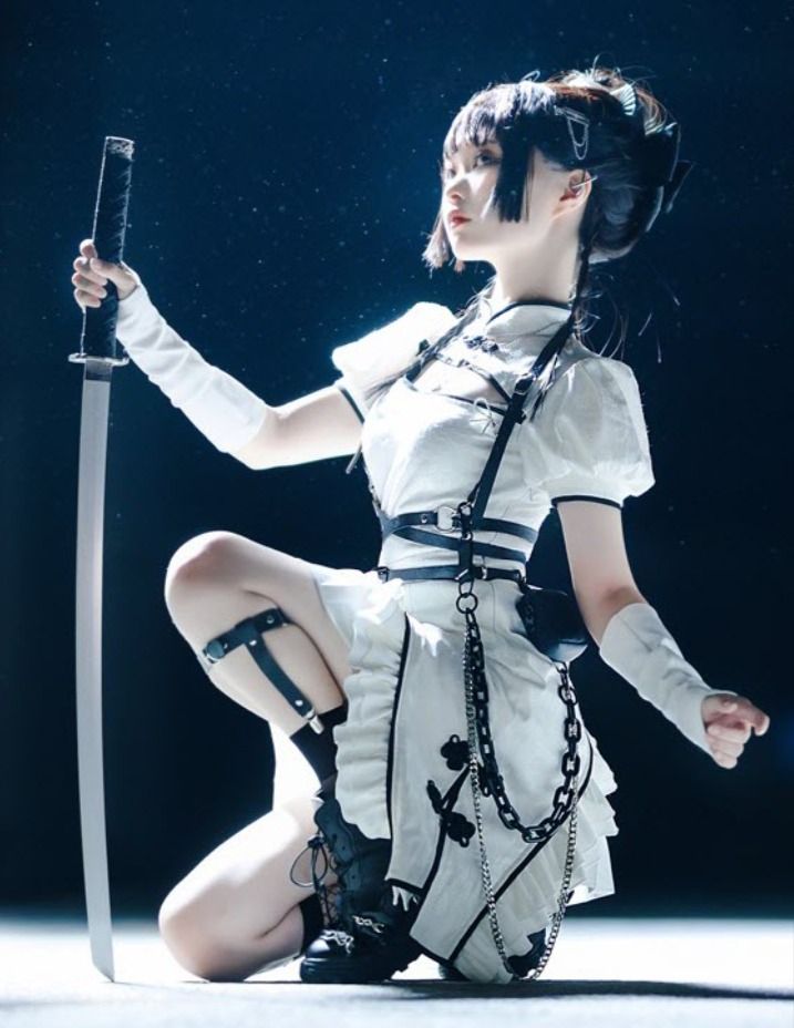 a young woman in a white dress and black boots kneels on the floor, holding a katana sword in her right hand. she is looking away from the camera with a serious expression on her face. the background is dark and blurry. the image has a dramatic and moody atmosphere.