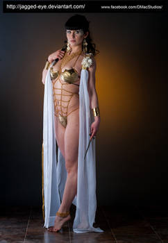 a fullbody shot of a woman posing in a studio setting. she is wearing a gold and white costume that resembles ancient greek or roman attire.  the costume includes a gold metallic bodice and chainlike details, partially sheer, over a white robe.  she holds a dagger in one hand. the background is dark, focusing attention on the model. her expression is serious and somewhat mysterious.  the overall style suggests a fantasy or mythological theme.