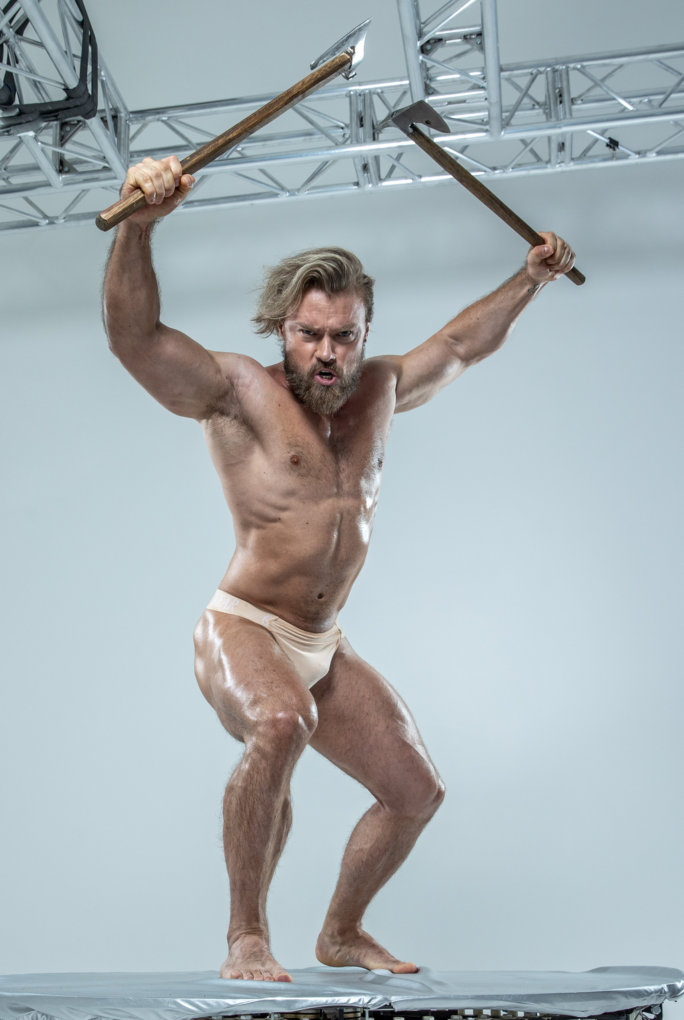 a muscular, barechested man with long blond hair and a beard stands on a platform in a photography studio.  he is oiled up and appears very strong.  he is holding axes in both hands above his head in a battle pose, his mouth open in a yell. his underwear is a peach color.  behind him is a white wall and part of a metal structure.