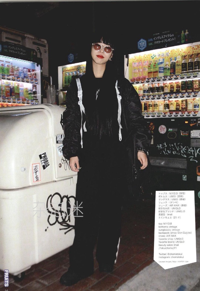 a young woman is posing in front of vending machines on a city street at night.  she is wearing a black bomber jacket, black pants with white stripes down the sides, a black shirt, and sunglasses.  she has on a black backpack with white straps.  her hair is dark and straight, and her makeup is simple.  the photo is taken from the front at eye level.  the lighting is bright and artificial.  the overall tone of the image is edgy and cool.
