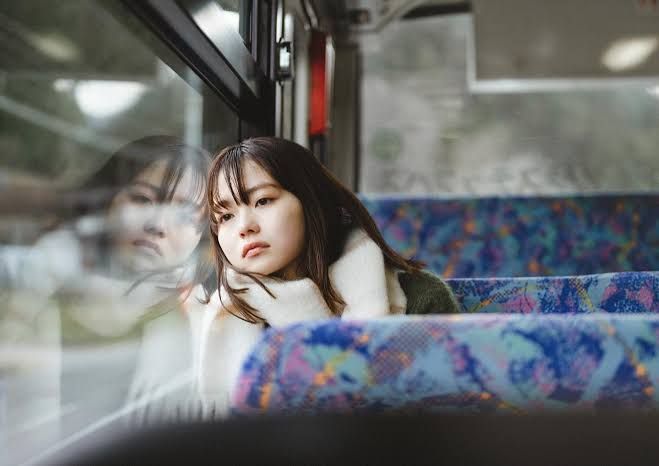 a young asian woman is sitting alone on a bus, looking out the window with a pensive expression. her reflection is visible in the window. she is wearing a sweater and scarf and appears sad or thoughtful. the bus seat has a blue and purple patterned fabric.
