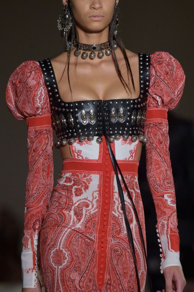a model is wearing a striking red and white paisley dress with a black studded leather bustierstyle top and choker. the dress has long puff sleeves and a cutout design at the waist. a row of buttons runs down the front of the skirt. she is also wearing ornate silver earrings.