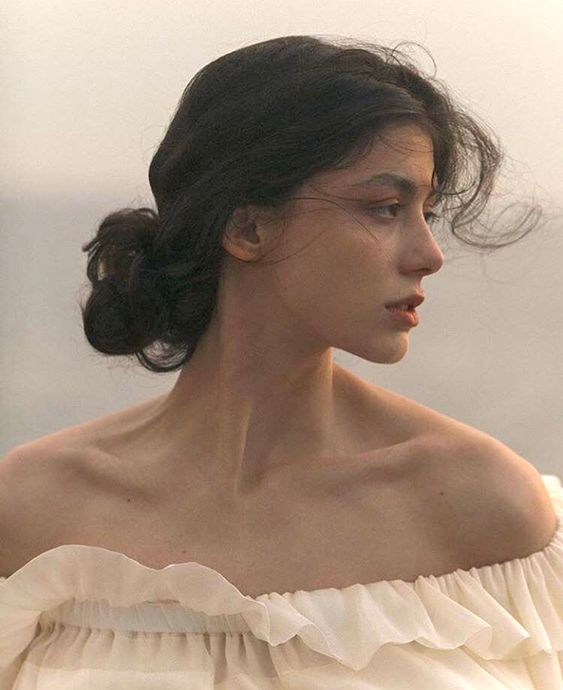 a young woman with dark hair pulled back in a loose bun is shown in a side profile view. she is wearing a white offtheshoulder blouse with ruffles and her expression is contemplative, gazing off into the distance. the lighting is soft and natural, giving the image a dreamy and romantic feel. the color palette consists of muted tones of beige, brown, and white, contributing to the vintage aesthetic of the photograph.