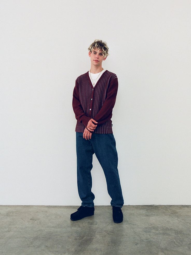 a young man with short blonde hair stands against a plain white background. he is modeling a burgundy and light purple striped cardigan, dark wash jeans, and darkcolored shoes. his pose is casual, hands clasped in front of him. the overall style is minimalist and contemporary.