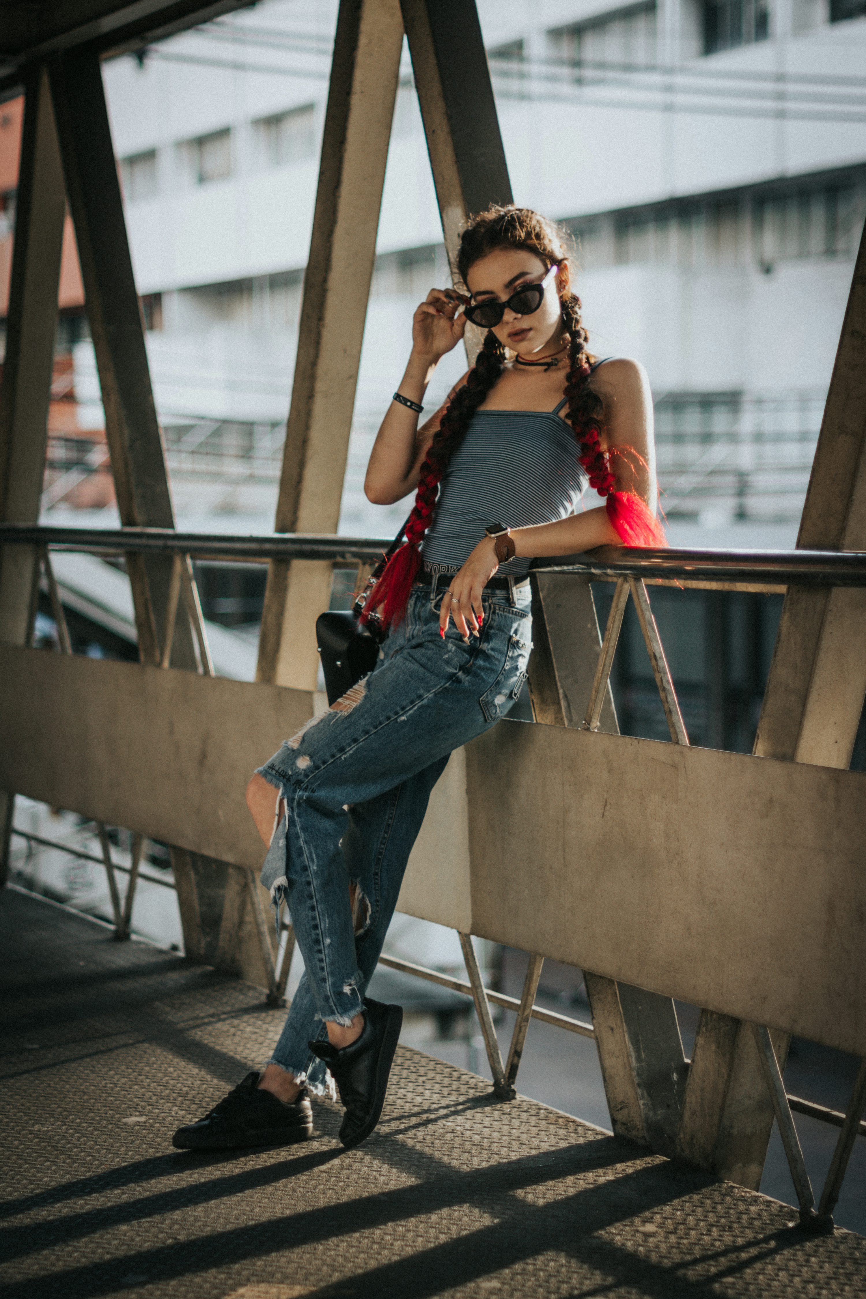 a young woman with redpigtailed hair is captured sitting on a metal railing of a city overpass. she's wearing ripped blue jeans, a gray tank top, black sneakers, and sunglasses.  she holds a small purse or handbag and her pose suggests a confident, stylish attitude. the setting is urban, with the city visible in the background.  strong shadows cast by the sun emphasize the texture of the metal structure and the woman's clothing.