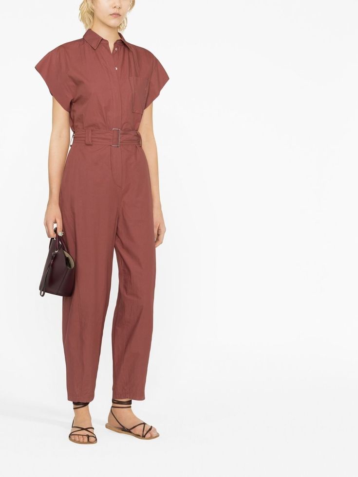 a woman wearing a red jumpsuit, a black bag, and sandals is standing against a white background. she is looking away from the camera.