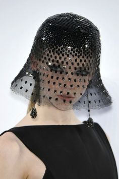 a woman is wearing a black hat with a black veil that covers her eyes. she is looking at the camera with a serious expression. the hat is made of a delicate material and has a lot of embellishments. the woman is wearing a simple black dress that is tailored to her body. she is wearing eyeliner and has long, dark eyelashes. she is looking at the camera with a serious expression. she is posing for a fashion photoshoot. the lighting is bright and wellbalanced.