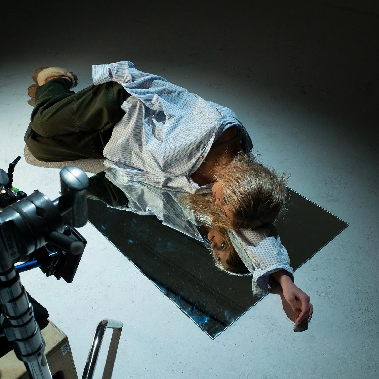 a person is lying on a square mirror in a studio setting. the person is wearing a striped blue and white shirt, olive green pants, and furlined slippers. their reflection is visible in the mirror. a film camera and lighting equipment are also visible in the shot, suggesting it's a photoshoot. the background is a plain white floor.