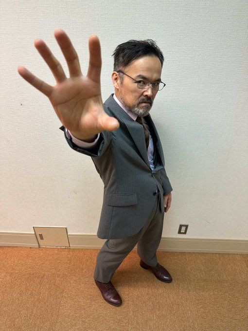 a middleaged asian man in a gray suit stands against a plain wall. he is wearing glasses and has a serious expression on his face. he is holding up his left hand in a "stop" gesture. the image is taken from a low angle, making the man appear taller and more imposing. the lighting is artificial and slightly overhead. the overall tone of the image is serious and businesslike.