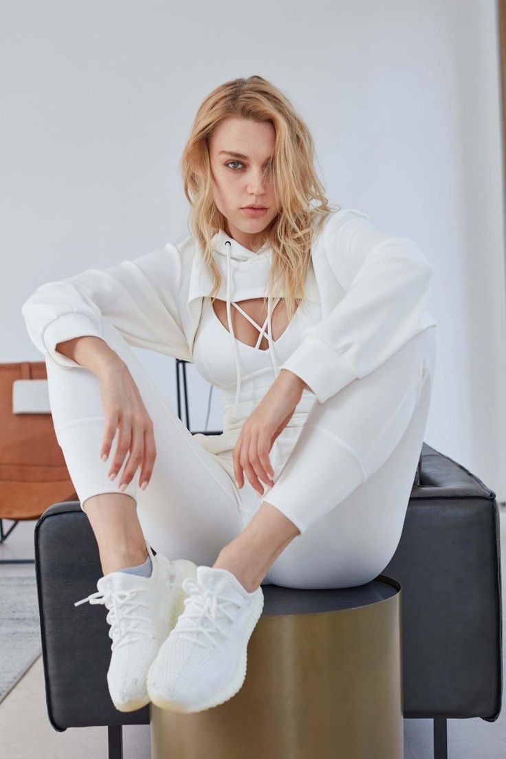 a blonde woman poses in a white athleisure outfit consisting of a cropped hoodie, sports bra, and leggings. she sits on a dark grey couch with her feet resting on a gold cylinder. the studio setting is minimally decorated with white walls and hints of brown furniture. the soft lighting illuminates the scene with a natural feel.