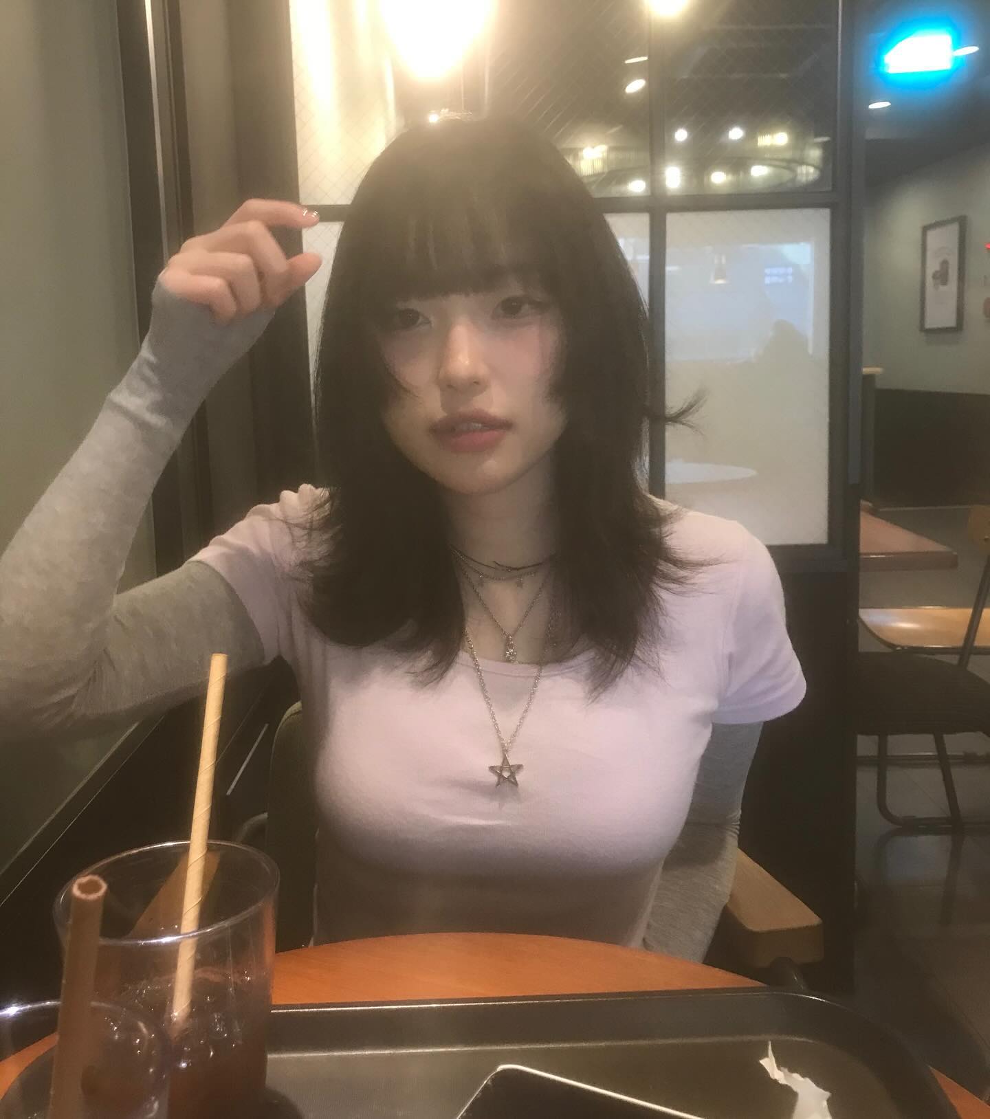 a young woman with dark hair and a fringe sits at a coffee shop table. she is wearing a light purple tshirt and has a necklace with a star pendant. she holds her hair with one hand while looking at the camera for a selfie. on the table in front of her is a glass of iced coffee with a straw and a cell phone. the cafe is lit with warm overhead lighting.