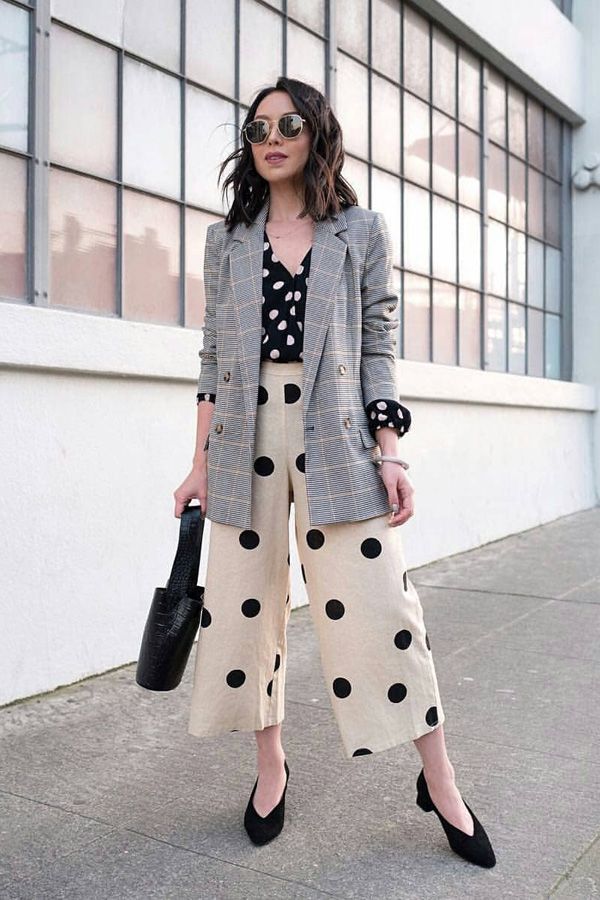 a stylish woman is standing outdoors in an urban setting. she's wearing a grey plaid blazer over a black polka dot blouse and beige polka dot culottes.  she's accessorized with black pointedtoe shoes, sunglasses, and a black handbag.  the overall look is a blend of casual and professional styles. the background features a modern building with large windows. the photo appears to be taken on a sunny day.