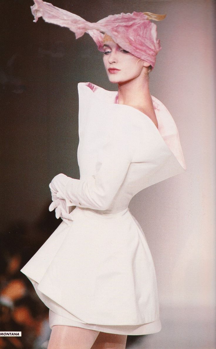 a fashion model is on a runway wearing a white dress, gloves, and a pink hat. the dress has an exaggerated collar and a flared skirt. the model is looking to the right. the brand "montana" is visible in the lowerleft corner of the image.