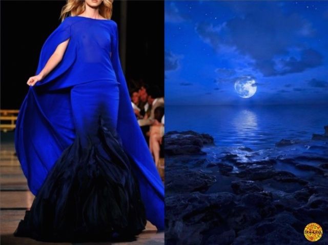 a beautiful woman in a long blue dress is walking down a runway. the dress has a capelike top and a mermaid skirt. she is walking towards the camera. the background is a beautiful night scene with a full moon over the ocean. there are rocks in the foreground. the woman's dress is the same color as the sky and the moon. this is a very elegant and sophisticated image.