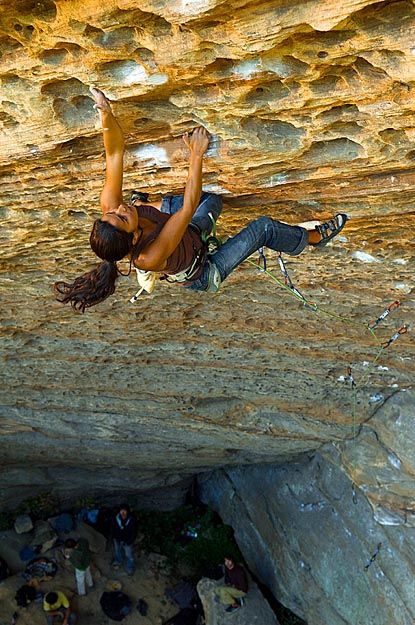 a female rock climber is shown midway up a steep rock face. she is seen from a low angle, with her body silhouetted against the bright sky. the texture of the rock is visible, as is the lush greenery at the base of the cliff. several other people are visible at the bottom, either climbing or observing.  the scene conveys a sense of adventure, skill, and the beauty of the natural world.